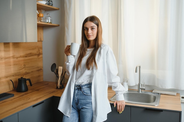 A young beautiful caucasian woman stands in the kitchen with a white cup of coffee or tea in the morning A lonely girl is enjoying a cup of fresh hot drink