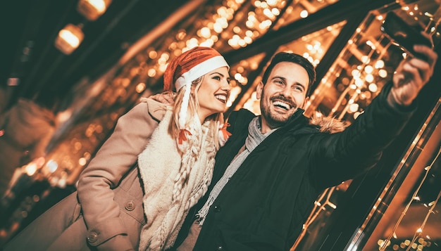 Young beautiful cheerful couple taking a selfie in the city street at new year's night with a lot of lights on background.