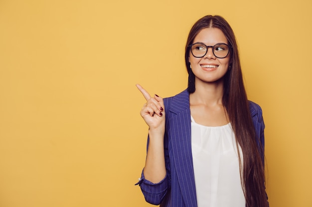 Young beautiful cheerful girl in dark blue jacket over yellow background smiling and looking at the camera pointing to the side at copy space.