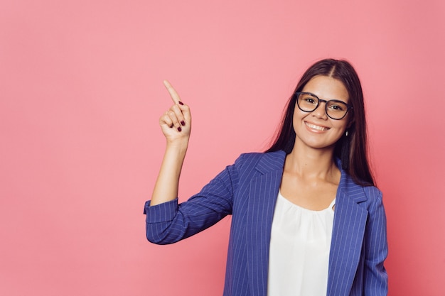 Young beautiful cheerfull girl in dark blue jacket over pink background smiling and looking at the camera pointing to the side at copy space.