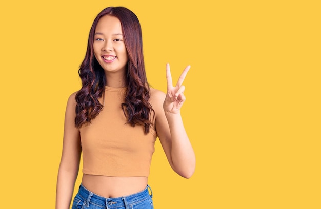 Young beautiful chinese girl wearing casual clothes smiling with happy face winking at the camera doing victory sign. number two.