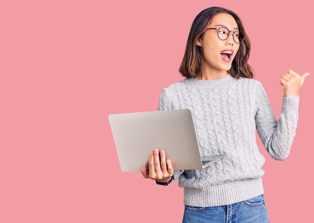 Young beautiful chinese girl wearing glasses holding laptop pointing thumb up to the side smiling happy with open mouth