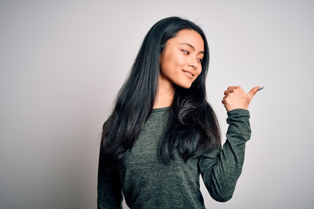 Young beautiful chinese woman wearing casual tshirt over isolated white background smiling with happy face looking and pointing to the side with thumb up