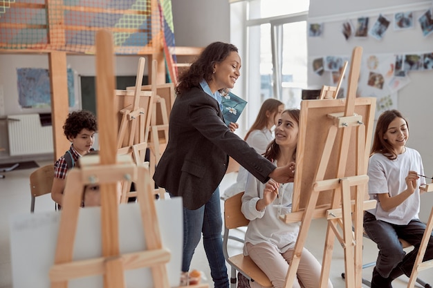 Young beautiful confident teacher is helping a kid to draw on a group lesson in a white modern minimalistic classroom