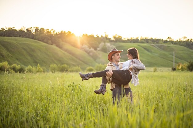 Young beautiful couple on a green meadow in the rays of the setting sun a man holds a girl in his arms cowboy clothesrustic style