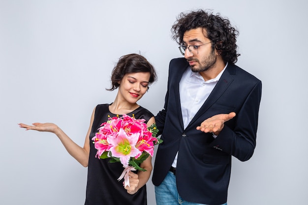 Young beautiful couple man and woman with bouquet of flowers looking confused raising arms celebrating valentine