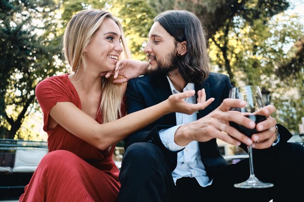 Young beautiful couple with glass of red wine happily looking at each other and talking on date in restaurant outdoor