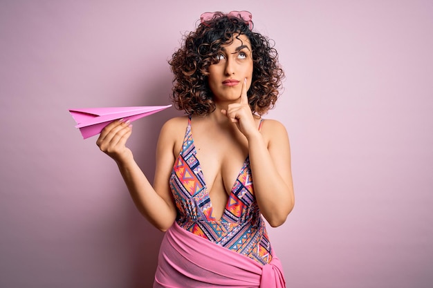 Young beautiful curly arab woman on vacation wearing swimsuit holding pink paper airplane serious face thinking about question very confused idea