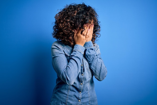 Young beautiful curly arab woman wearing casual denim shirt standing over blue background with sad expression covering face with hands while crying Depression concept