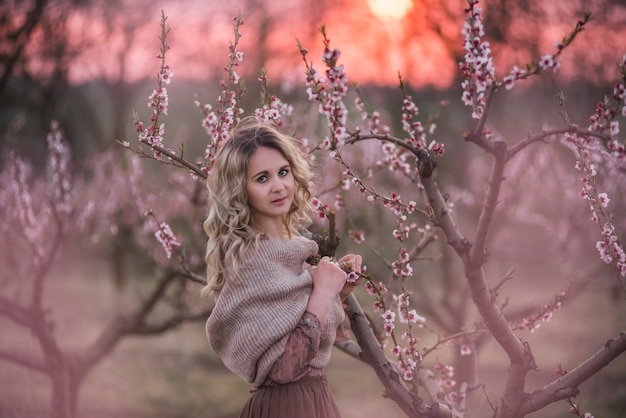 Young beautiful curly blonde woman in brown pleated skirt pink blouse covered shoulders with knitted scarf, stands in blooming peach sunset gardens