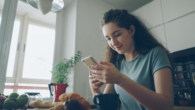 Young beautiful curly pretty caucasian woman sitting at table in nice kitchen using smartphone she is texting someone and smiling calm and happy