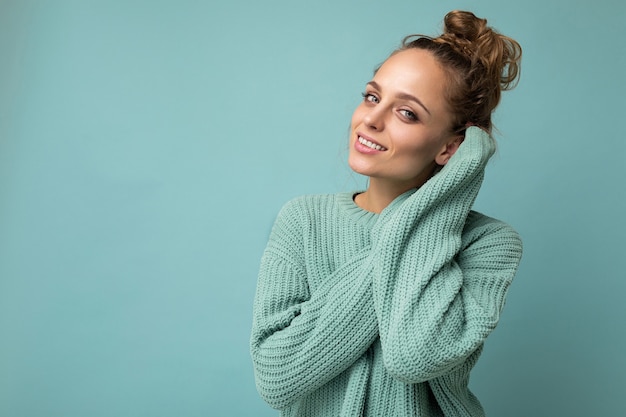 Young beautiful cute curly blonde woman with sexy expression, cheerful and happy face wearing trendy blue sweater isolated over blue background with copy space