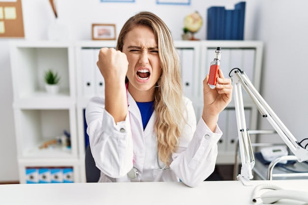 Young beautiful doctor woman holding electronic cigarette at the clinic annoyed and frustrated shouting with anger yelling crazy with anger and hand raised