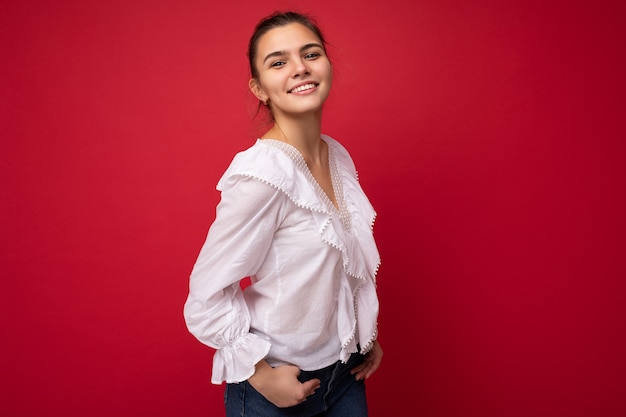 Young beautiful european stylish brunette woman wearing white blouse isolated over red background with positive sincere emotions. Simple and natural looking at the camera. Free space.