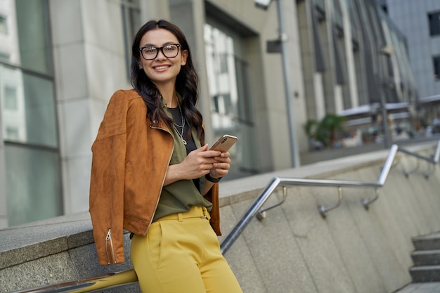 Young beautiful and fashionable caucasian woman holding her smartphone looking at camera and smiling