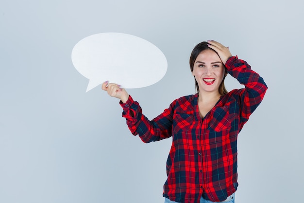 Young beautiful female in casual shirt, jeans holding speech bubble while keeping hand on head and looking blissful , front view.