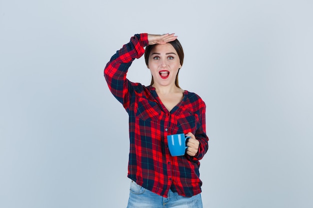 Young beautiful female in casual shirt, jeans keeping hand over head while holding cup and looking joyful , front view.