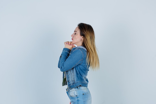 Young beautiful female in denim outfit holding clasped hands in pleading gesture and looking hopeful .