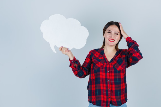 Young beautiful female keeping hand on head while holding speech bubble in casual shirt, jeans and looking cheerful. front view.