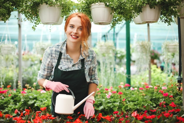 Young beautiful florist in apron standing with watering can and happily looking in camera while working in greenhouse