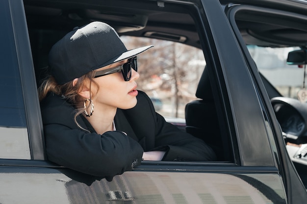 A young beautiful girl in a black car looks out of the window. A stylish girl in glasses and a cap rides in a car, leaning out of the window.