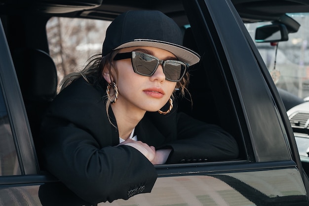 A young beautiful girl in a black car looks out of the window. A stylish girl in glasses and a cap rides in a car, leaning out of the window.