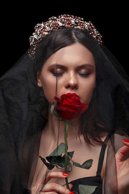 Young beautiful girl in a black headband and black veil in a corset with a scarlet rose in her hands on a black isolated background