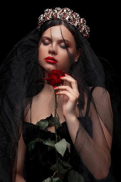 Young beautiful girl in a black headband and black veil in a corset with a scarlet rose in her hands on a black isolated background