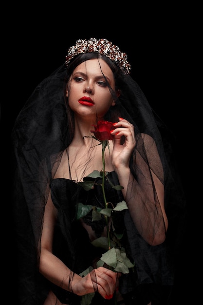 Young beautiful girl in a black headband and black veil in a corset with a scarlet rose in her hands on a black isolated background