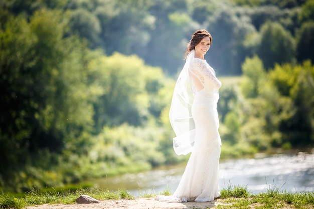 Young beautiful girl bride in white dress stay on coast of wide river and waits for the groom The bride at a high cliff near the river The best day of young woman