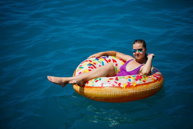 A young beautiful girl in a bright swimsuit lies on a large inflatable ring and floats on the blue sea on a bright sunny summer day