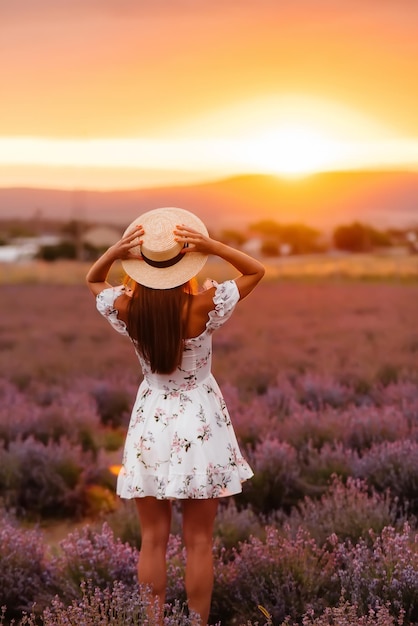 A young beautiful girl in a delicate dress and hat walks through a beautiful field of lavender and enjoys the aroma of flowers. Vacation and beautiful nature.