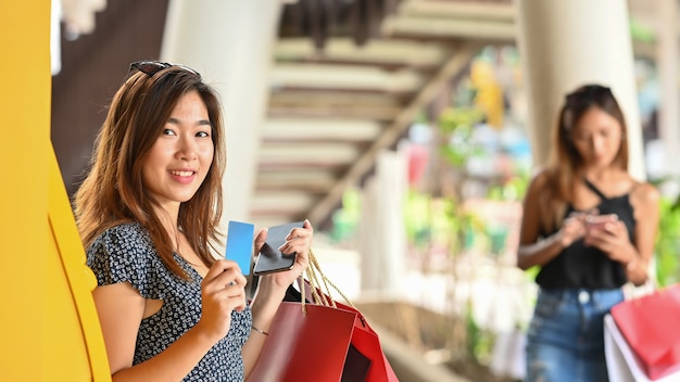 Young beautiful girl in dress holding a credit card and shopping bags in her hands while standing next to the yellow auto teller machine with blurred woman and supermarket 