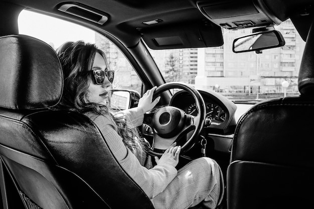 Young, beautiful girl driving a car. A stylish girl in a suit and glasses driving a black car. Black and white photo