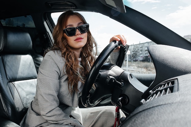 Young, beautiful girl driving a car. A stylish girl in a suit and glasses driving a black car.