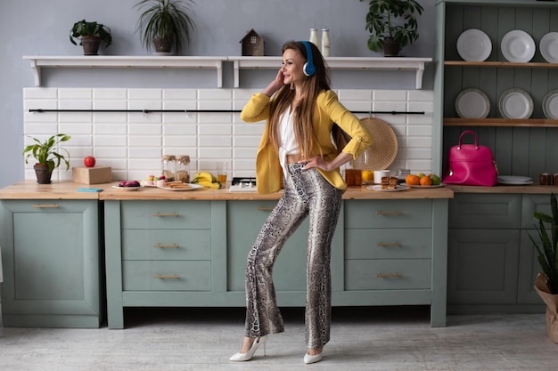 Young beautiful girl in fashionable clothes getting fun on kitchen Happy brunette model with long hair listening to music and dancing Stylish woman in yellow jacket and blue headphones singing