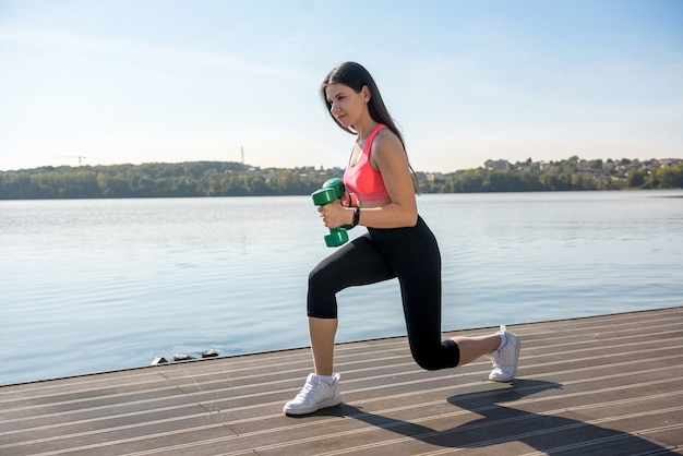 Young beautiful girl is engaged in morning gymnastics with dumbbells at the lake