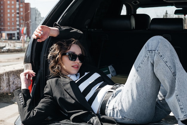 The young, beautiful girl is lying in the trunk of her black car. A stylish girl in glasses and a suit.