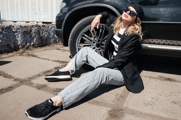 A young, beautiful girl is sitting on the asphalt near her black car in a black jacket, cap and glasses.