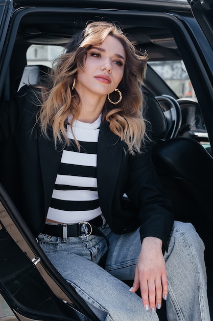 A young, beautiful girl is sitting in the back seat of a car. A stylish girl in a suit gets out of the car.