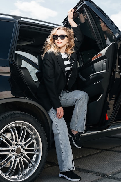 A young, beautiful girl is sitting in the back seat of a car. A stylish girl in a suit and glasses gets out of the car.