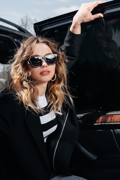 A young, beautiful girl is sitting in the back seat of a car. A stylish girl in a suit and glasses gets out of the car.