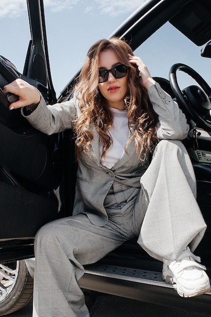 A young, beautiful girl is sitting in the driver's seat. A stylish girl in a suit and glasses opens the car doors