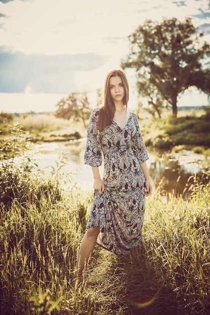 Young beautiful girl in a long dress standing against the sky and nature during the sunset