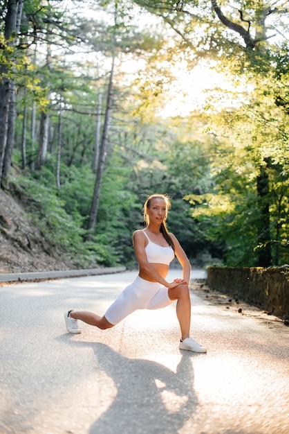 A young beautiful girl makes lunges and warm-up before running training, on the road in a dense forest, during sunset. A healthy lifestyle and running in the fresh air.