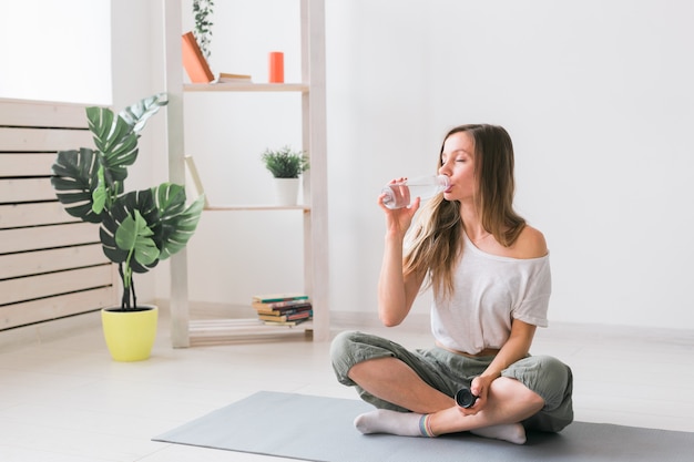 Young beautiful girl practicing yoga at mat, drinking water while break. Fitness and healthy lifestyle concept.