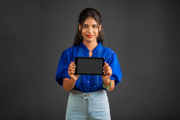 Young beautiful girl showing a blank screen of a smartphone or mobile or tablet phone on a grey background