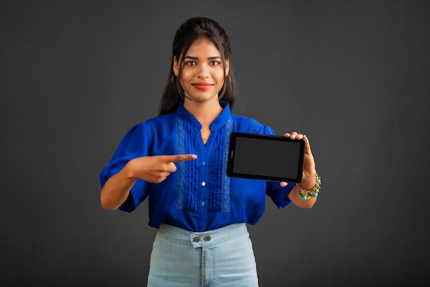 Young beautiful girl showing a blank screen of a smartphone or mobile or tablet phone on a grey background