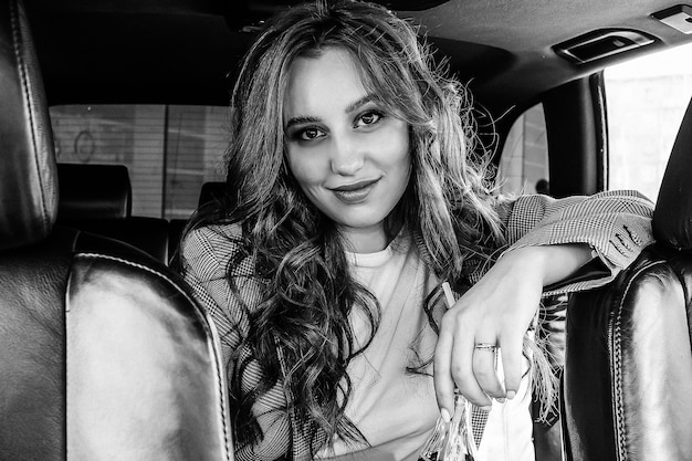 Young beautiful girl sitting in a car. A stylish girl in a suit drinks lemonade in a black car interior. Black and white photo.