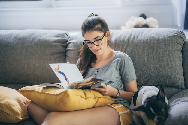 Young beautiful girl sitting on the sofa, reading a novel book and her dog stay by her side, lifestyle concept, hobby at home, a women wearing grey t-shirt and glasses, relax and chill